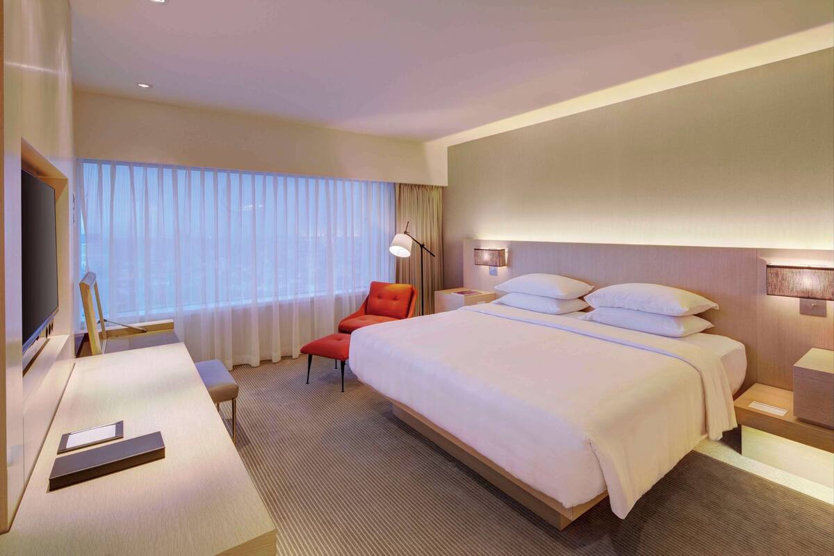 Une chambre d’hôtel avec un grand lit, un bureau avec une chaise, une télévision et une fenêtre avec des rideaux légers.