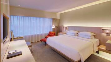 Ein Hotelzimmer mit einem großen Bett, einem Schreibtisch mit Stuhl, einem Fernseher und einem Fenster mit durchsichtigen Vorhängen.