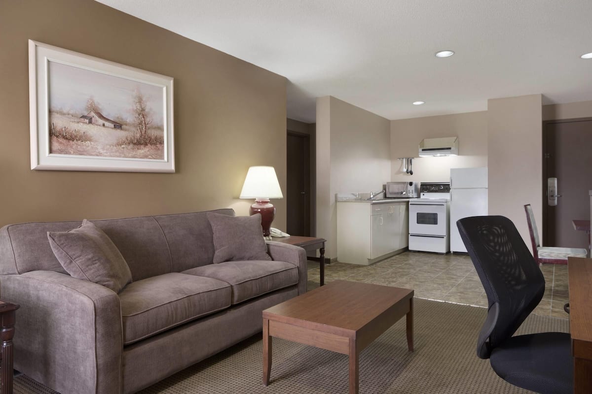 A living room with a sofa, a coffee table, a painting, a lamp, and a kitchen in the background.