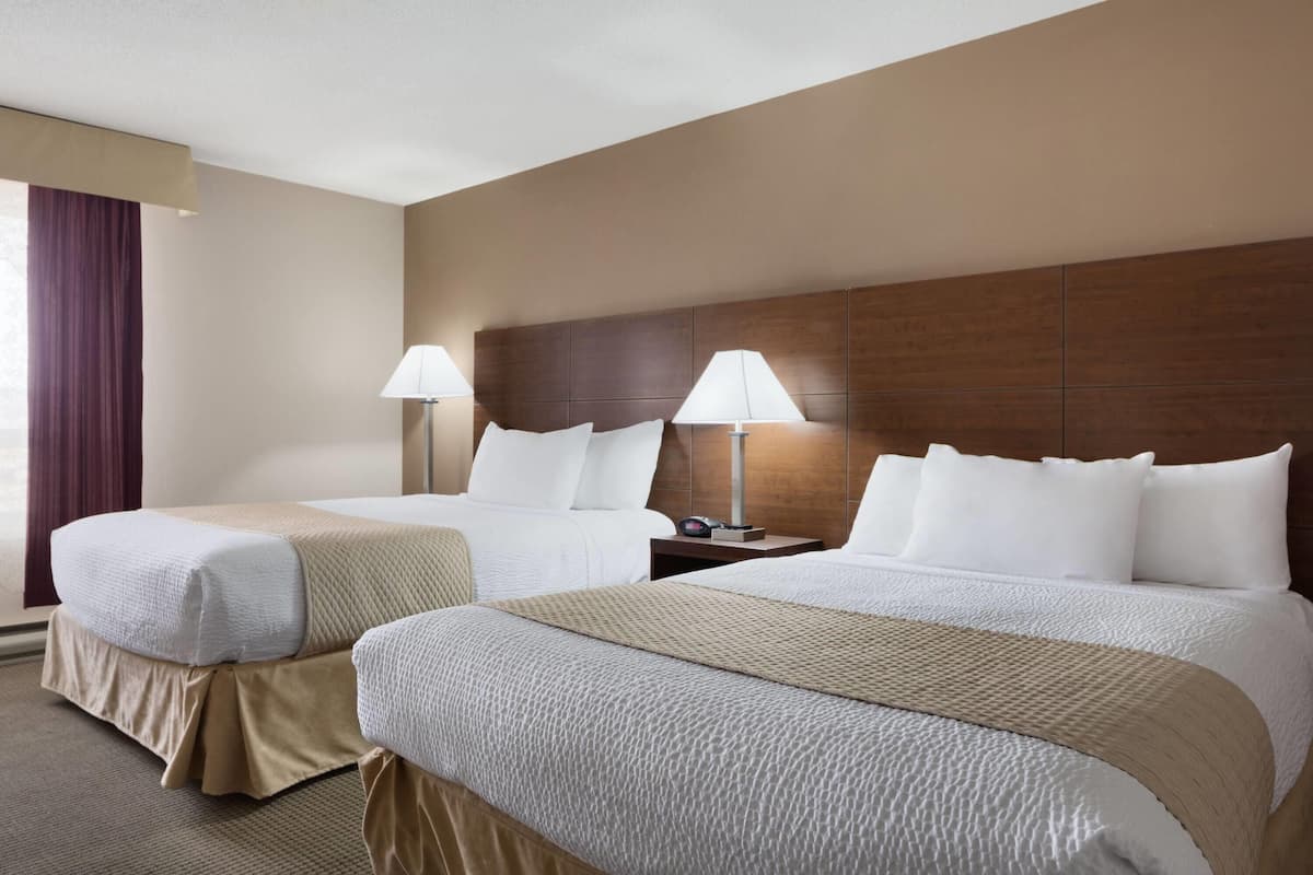 A hotel room with two beds, each with white linens and a beige accent. There are two bedside tables with lamps. The room has a neutral color scheme with beige walls and a brown headboard.