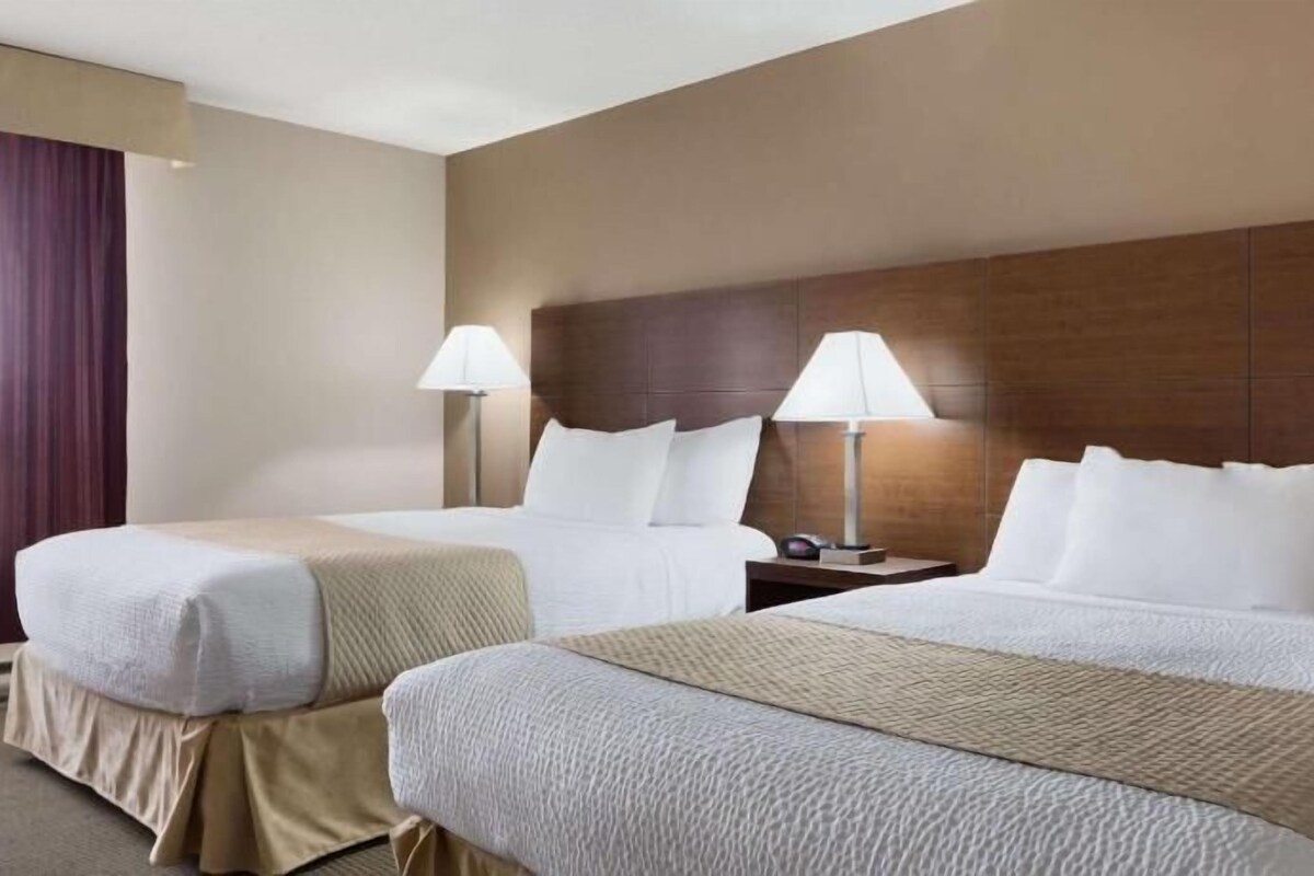 Une chambre d’hôtel avec deux lits, une tête de lit en bois, des lampes de chevet et une fenêtre avec des rideaux.