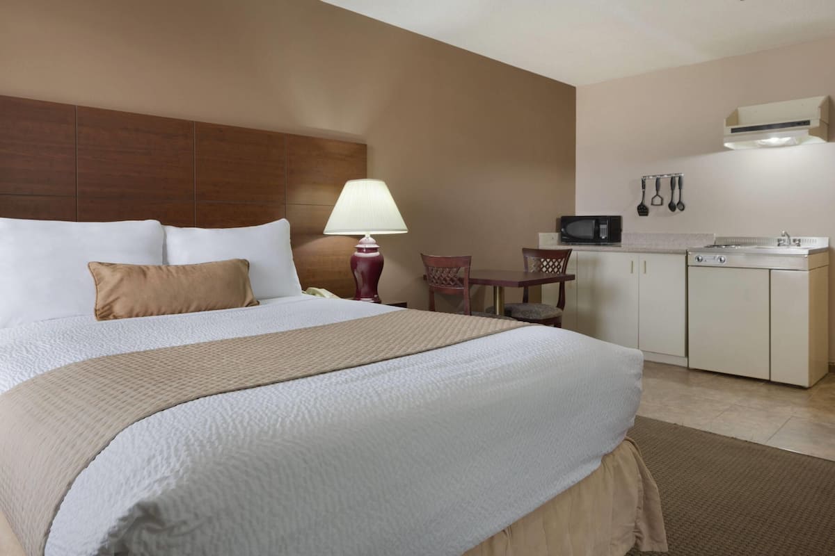 A hotel room with a large bed, a bedside table with a lamp, a small dining area, and a kitchenette.