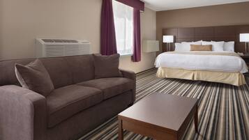 A hotel room with a brown sofa, a wooden coffee table, a striped carpet, a bed with white bedding, and an air conditioning unit.