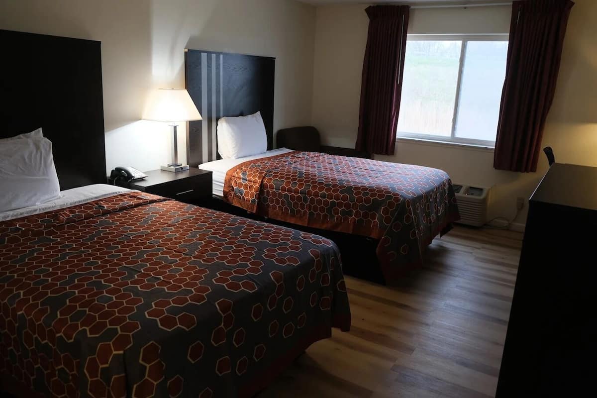 A hotel room with two beds, a desk with a lamp, a chair, and a window with curtains.
