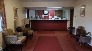 Zona de recepción con alfombra roja, sillas estampadas, un reloj y un mostrador con carteles informativos.