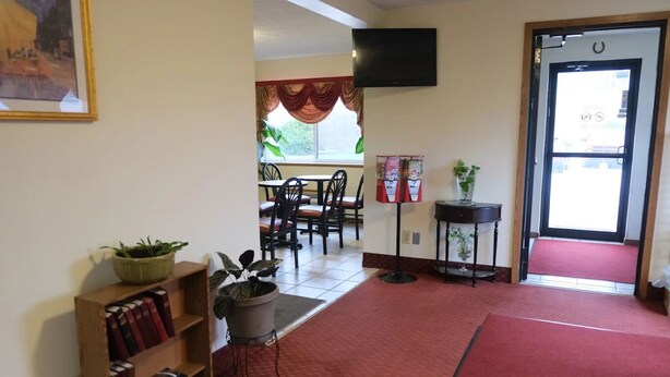 Un vestíbulo de hotel con alfombra roja, una estantería llena de libros, una planta en maceta, una mesa con sillas, una mesita con un florero y una puerta con felpudo rojo.