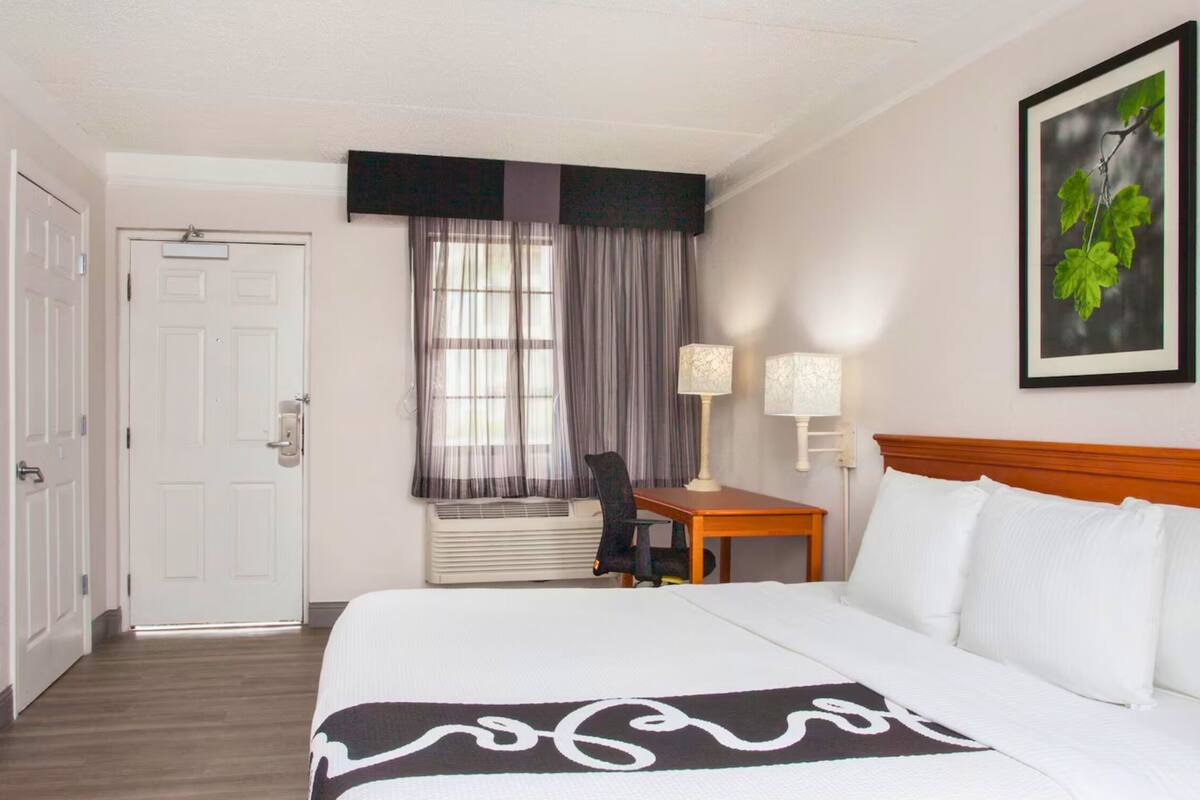 A hotel room with a bed, a desk with a chair, a television, and a window with curtains.