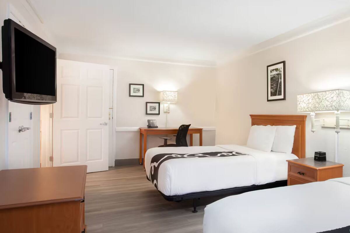A hotel room with two beds, a desk, a chair, a TV, and a lamp.