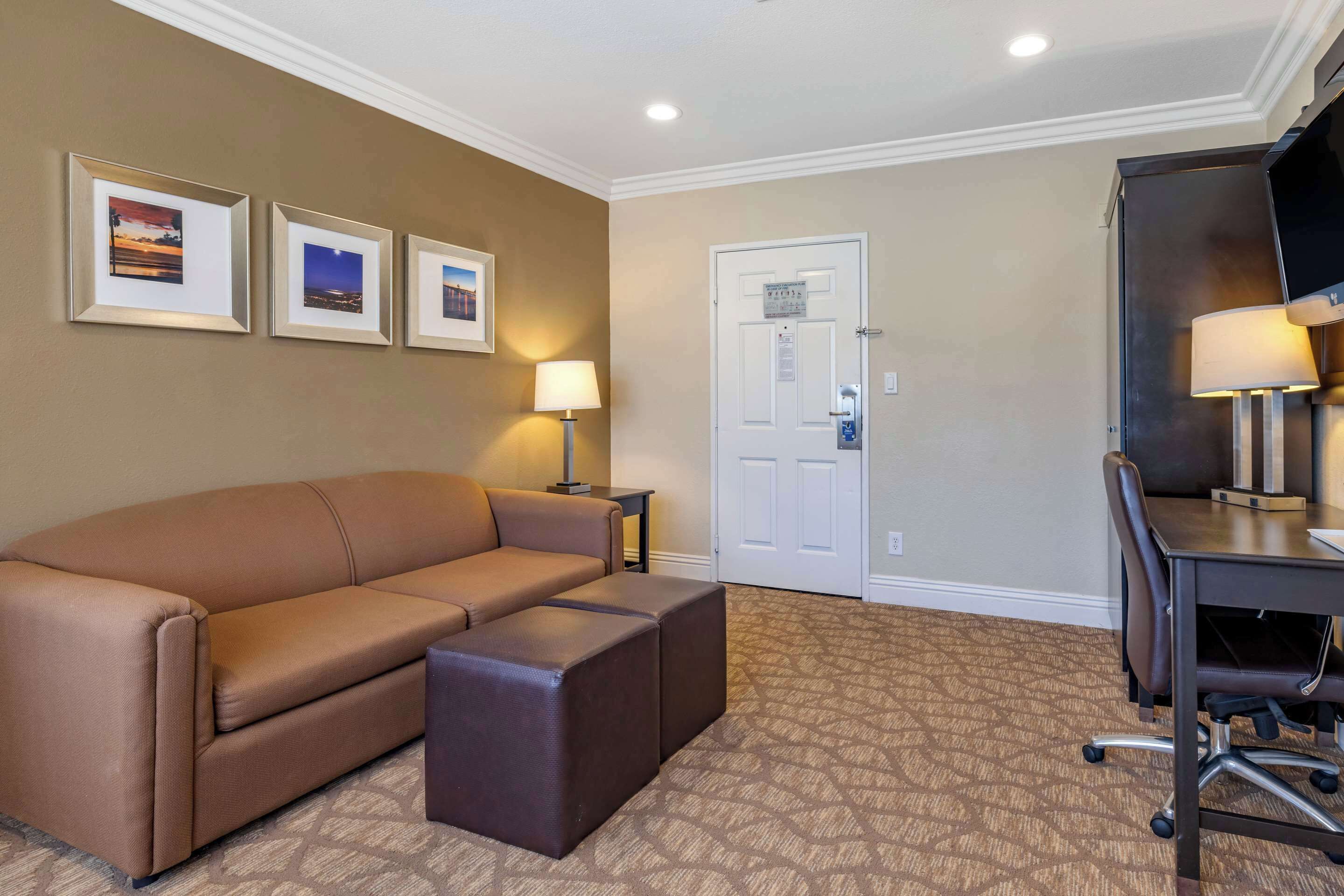 A hotel room with a brown sofa, a desk with a chair, a lamp, and framed pictures on the wall.