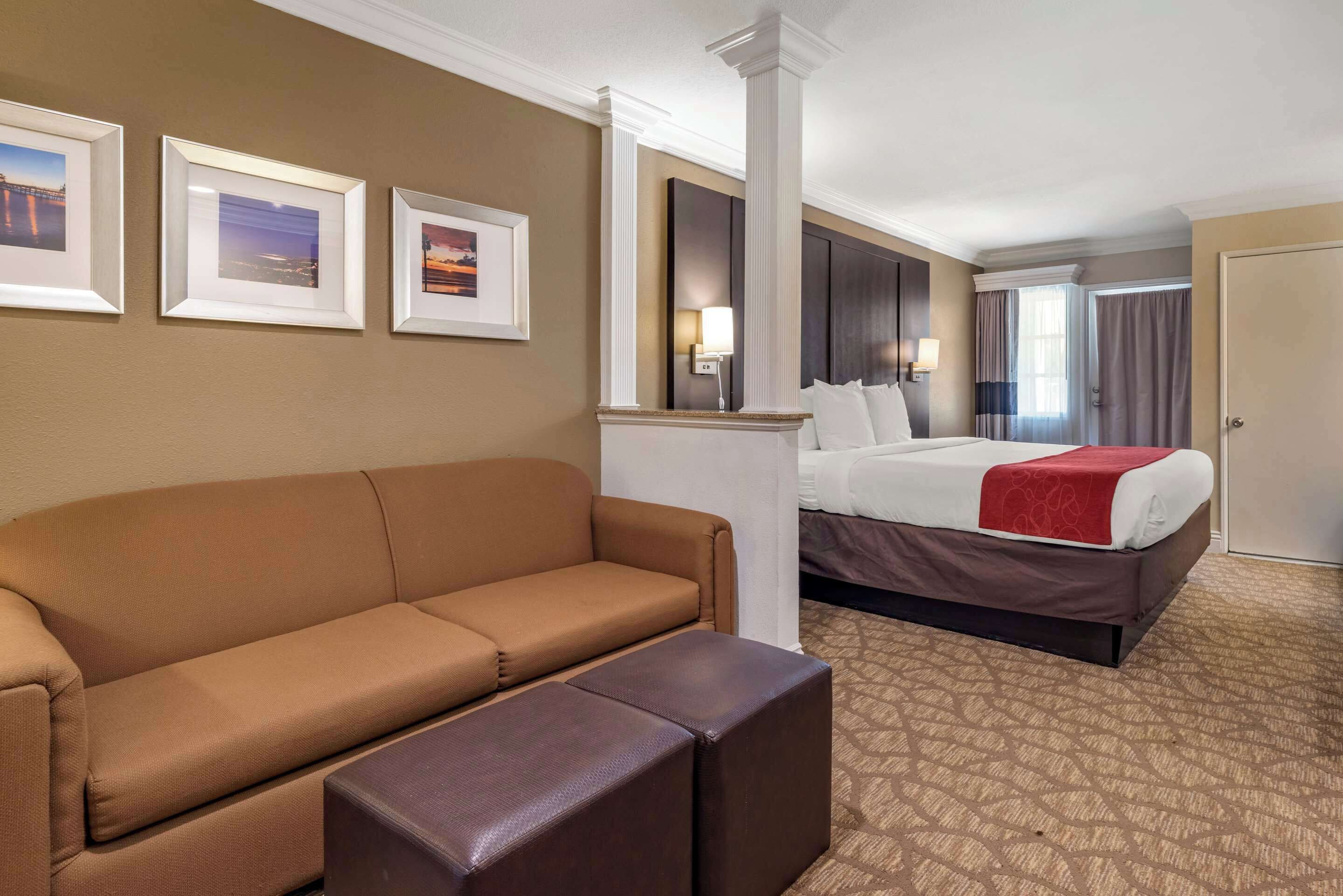 A hotel room with a brown sofa, a bed with a red blanket, and three framed pictures on the wall.