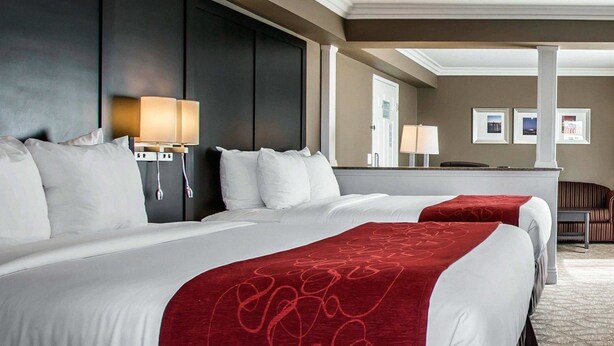 A hotel room with two beds, a dark headboard, bedside lamps, a red throw blanket, and a view of the hallway.