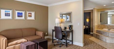 A hotel room with a beige sofa, a desk with a chair, a large mirror, and a bathroom with a bathtub and sink.