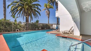 A swimming pool with a tiled edge, surrounded by palm trees and a white building with a balcony.