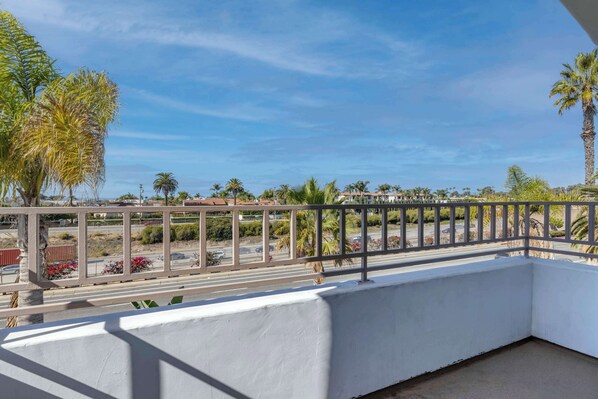 A balcony with a white railing overlooking a street with palm trees and a clear blue sky.