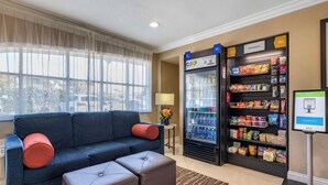 A hotel lobby with a sofa, ottoman, and a beverage refrigerator stocked with snacks and drinks.