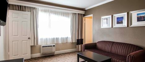 Une chambre d’hôtel avec une moquette à motifs, un canapé sombre, une petite table, une porte blanche, une fenêtre avec des rideaux et trois tableaux encadrés accrochés au mur.
