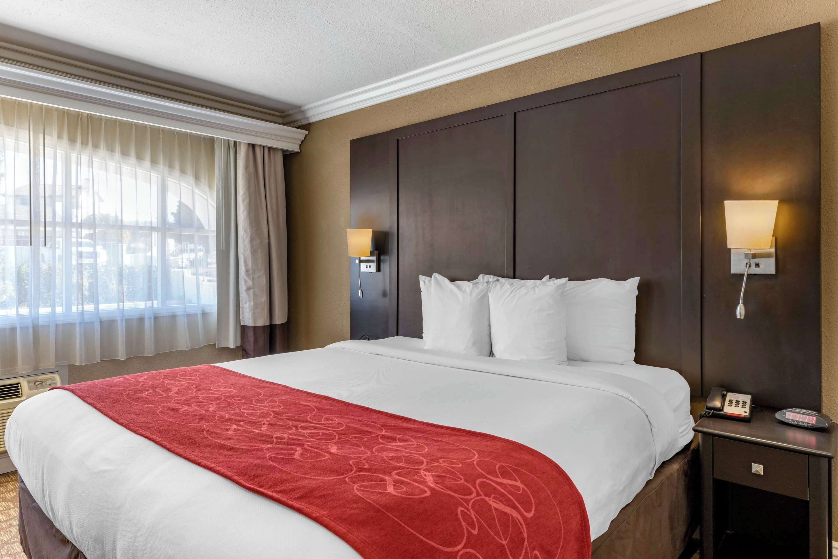 Een hotelkamer met een groot bed, een rode plaid, een donker hoofdeinde, twee bedlampen, een nachtkastje met een telefoon en een raam met gordijnen.
