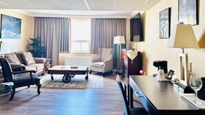 A hotel room with a living area featuring a sofa, armchairs, a coffee table, and a dining table with a lamp. There is a window with curtains, a plant, and artwork on the walls. - Travelodge by Wyndham Prince Albert (Prince Albert)
