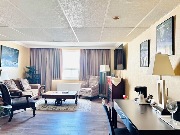 A hotel room with a living area featuring a sofa, armchairs, a coffee table, and a dining table with a lamp. There is a window with curtains, a plant, and artwork on the walls.