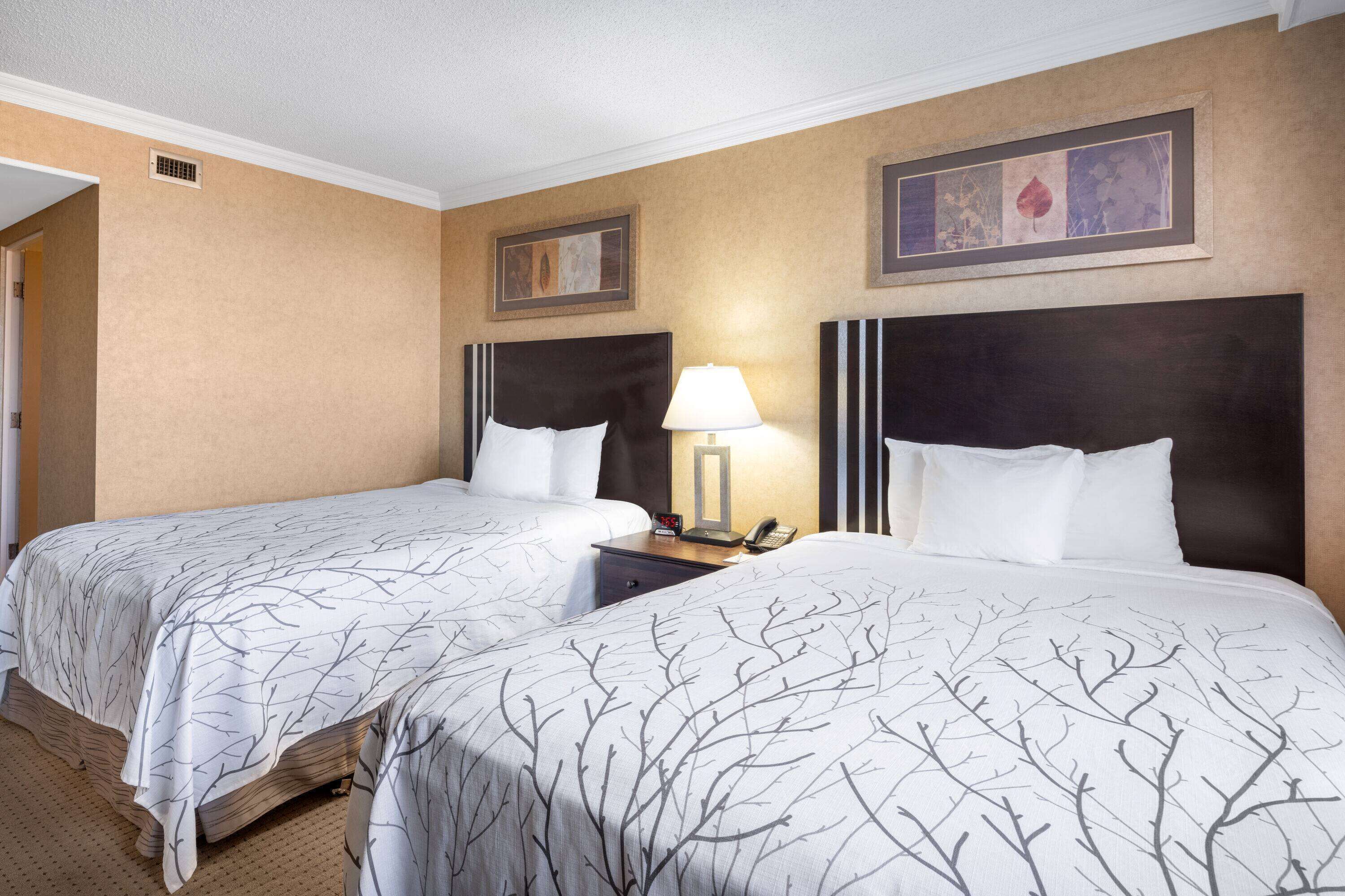 A hotel room with two beds, each with a dark headboard and white bedding. There is a nightstand with a lamp and a phone between the beds. The walls are beige, and there is a framed picture above the headboard on the right.