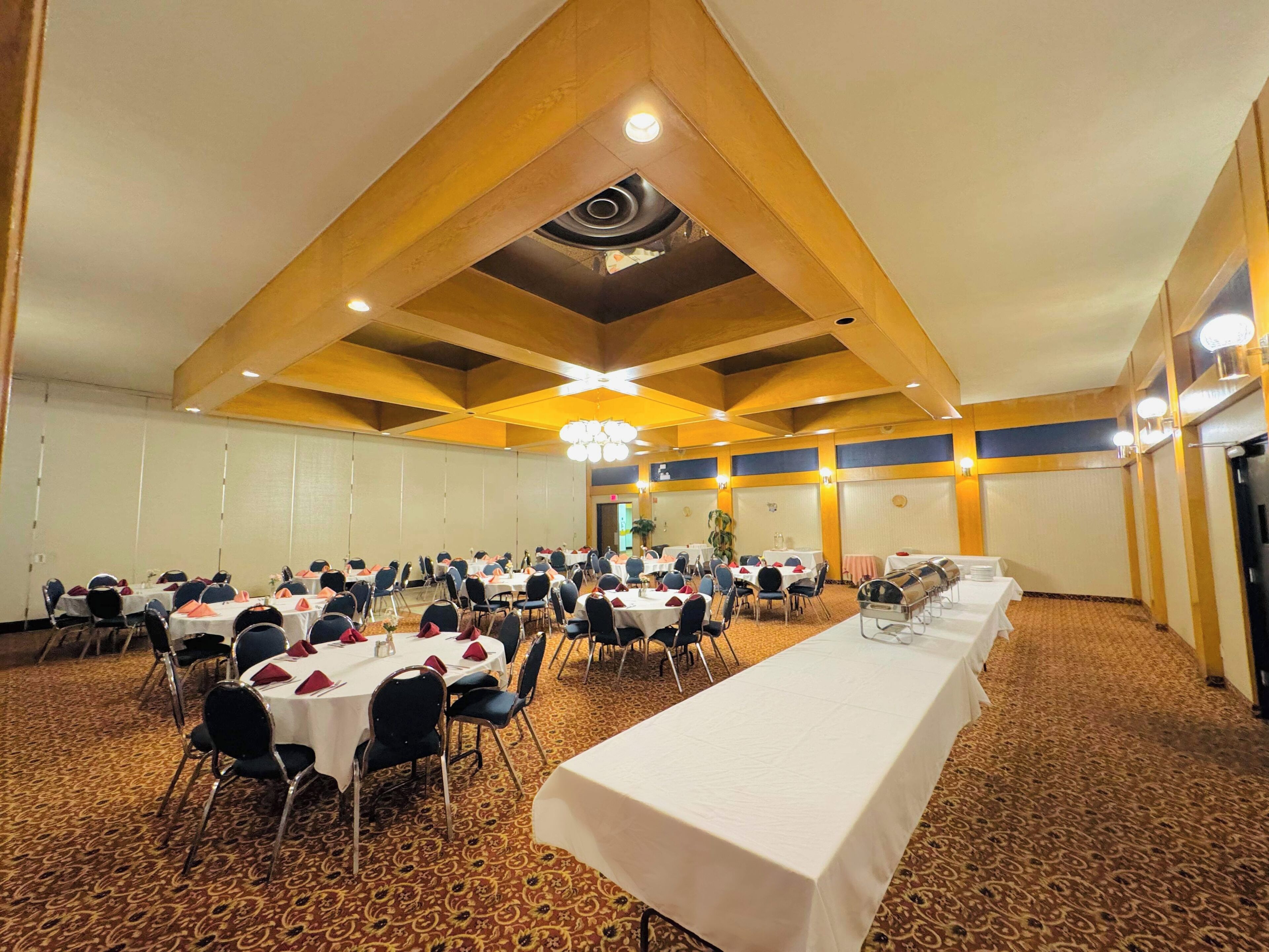 Een grote congreszaal met meerdere ronde tafels, elk gedekt met witte tafelkleden en rode servetten. De zaal heeft een hoge plafond met houten balken en inbouwverlichting.