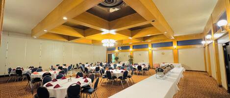 Een grote congreszaal met meerdere ronde tafels, elk gedekt met witte tafelkleden en rode servetten. De zaal heeft een hoge plafond met houten balken en inbouwverlichting.