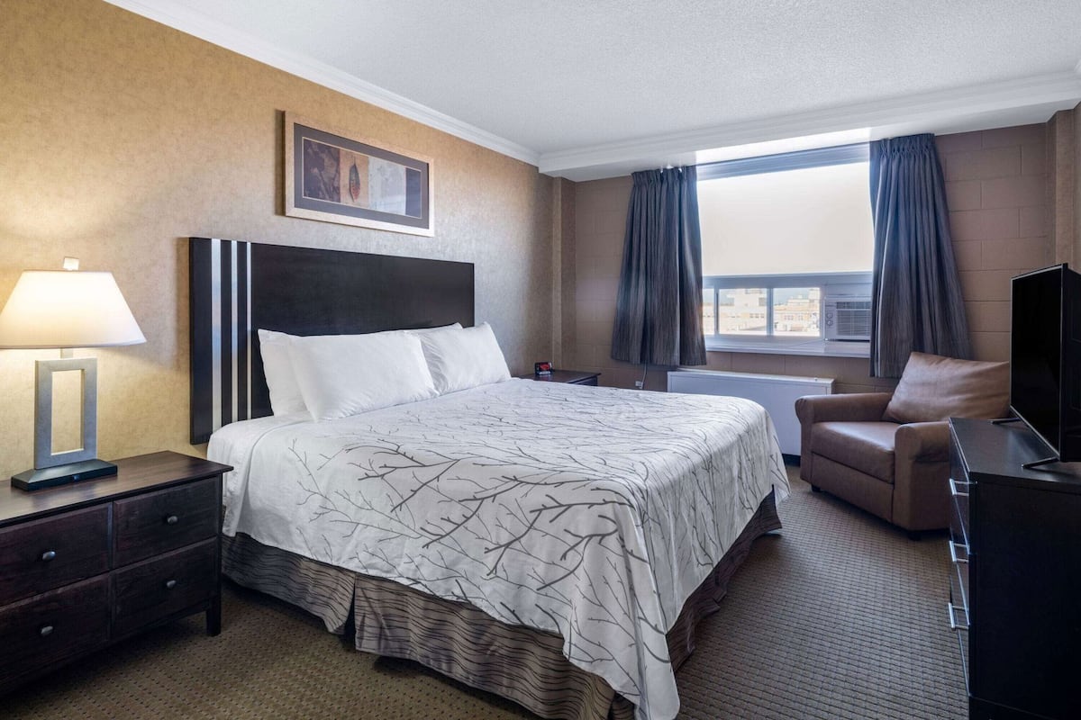 Une chambre d’hôtel avec un lit, un bureau avec une lampe, une chaise, une télévision et une fenêtre avec des rideaux.