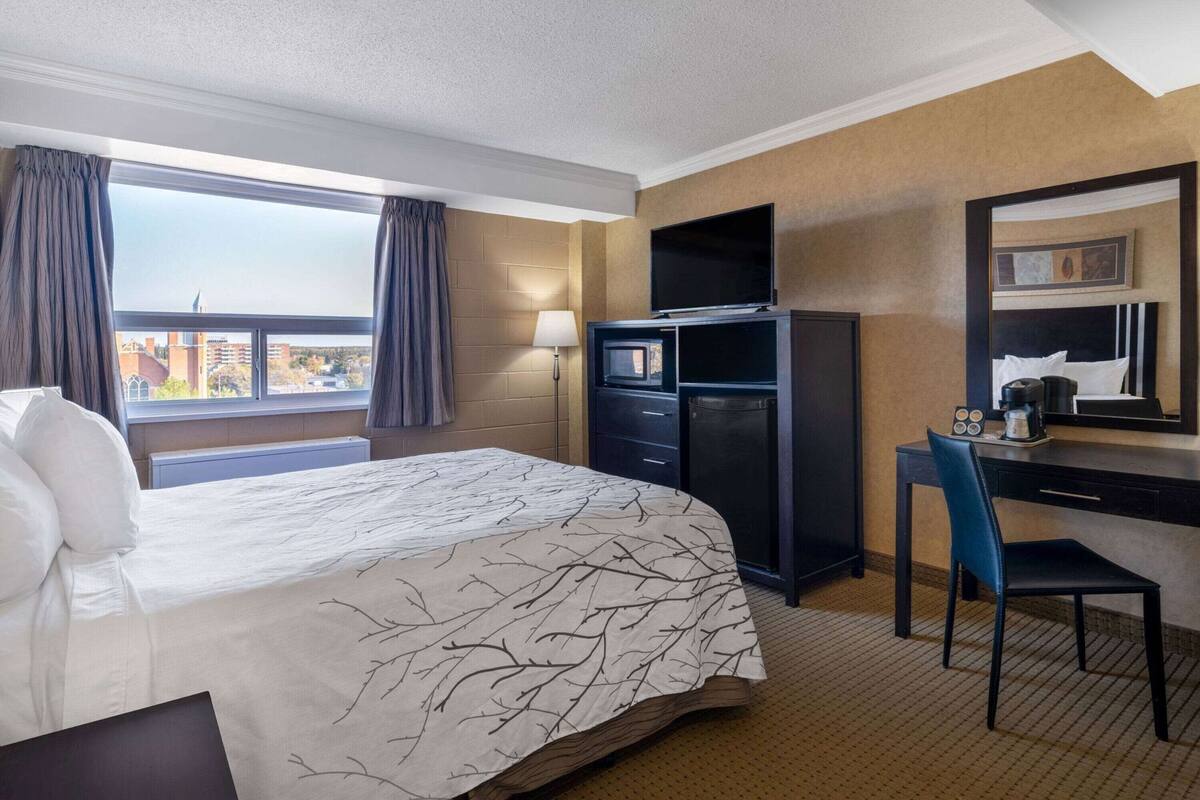 Une chambre d’hôtel avec un grand lit, un bureau avec une chaise, une télévision, un réfrigérateur et une fenêtre avec des rideaux.