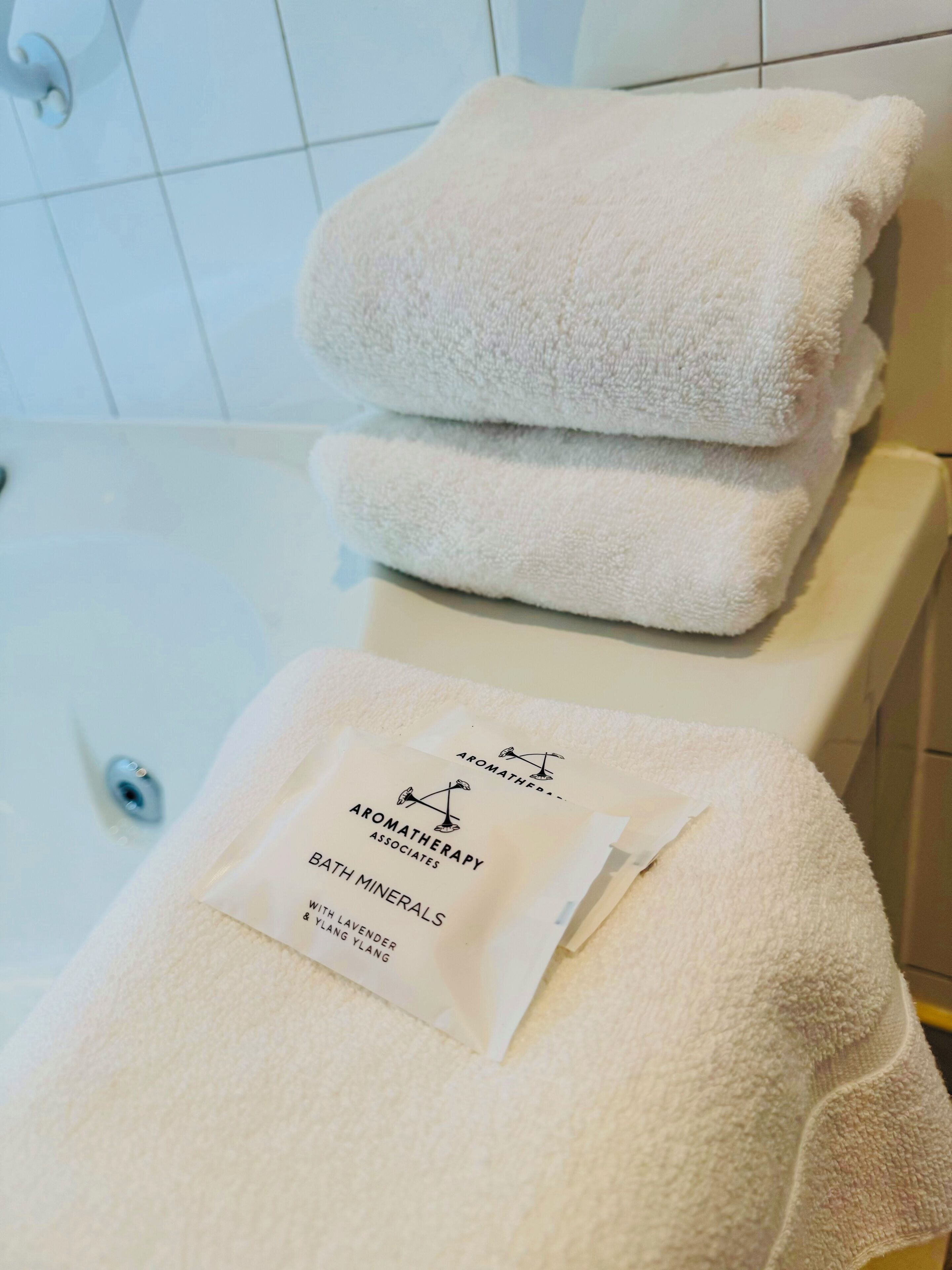 Une salle de bain avec une baignoire blanche, deux serviettes pliées et un sachet de sels de bain.