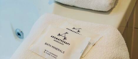 Une salle de bain avec une baignoire blanche, deux serviettes pliées et un sachet de sels de bain.