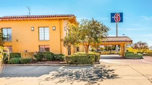 A yellow motel with a red-tiled roof, a sign indicating it's number 6, and a well-maintained front yard with hedges and a tree.