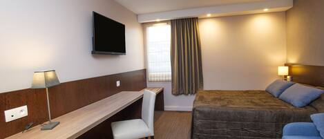 Une chambre d’hôtel avec un lit, un bureau, une chaise, une télévision et une fenêtre avec des rideaux.