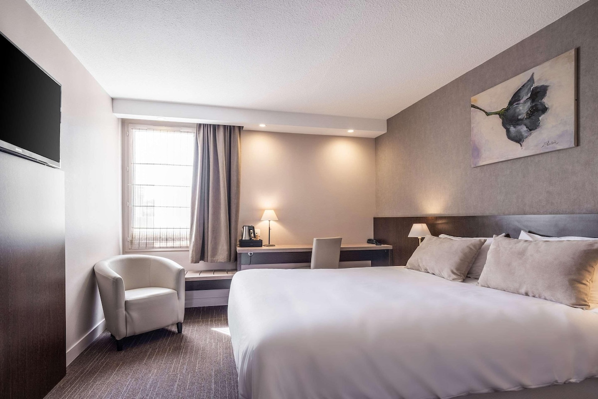Ein Hotelzimmer mit einem großen Bett, einem Schreibtisch mit Stuhl, einem Fernseher und einem Fenster mit Vorhängen.