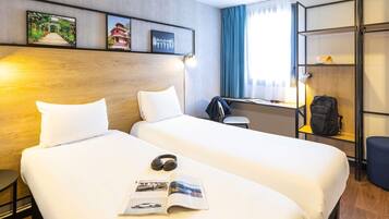 A hotel room with two beds, a desk, a chair, a bookshelf, and a window with curtains.