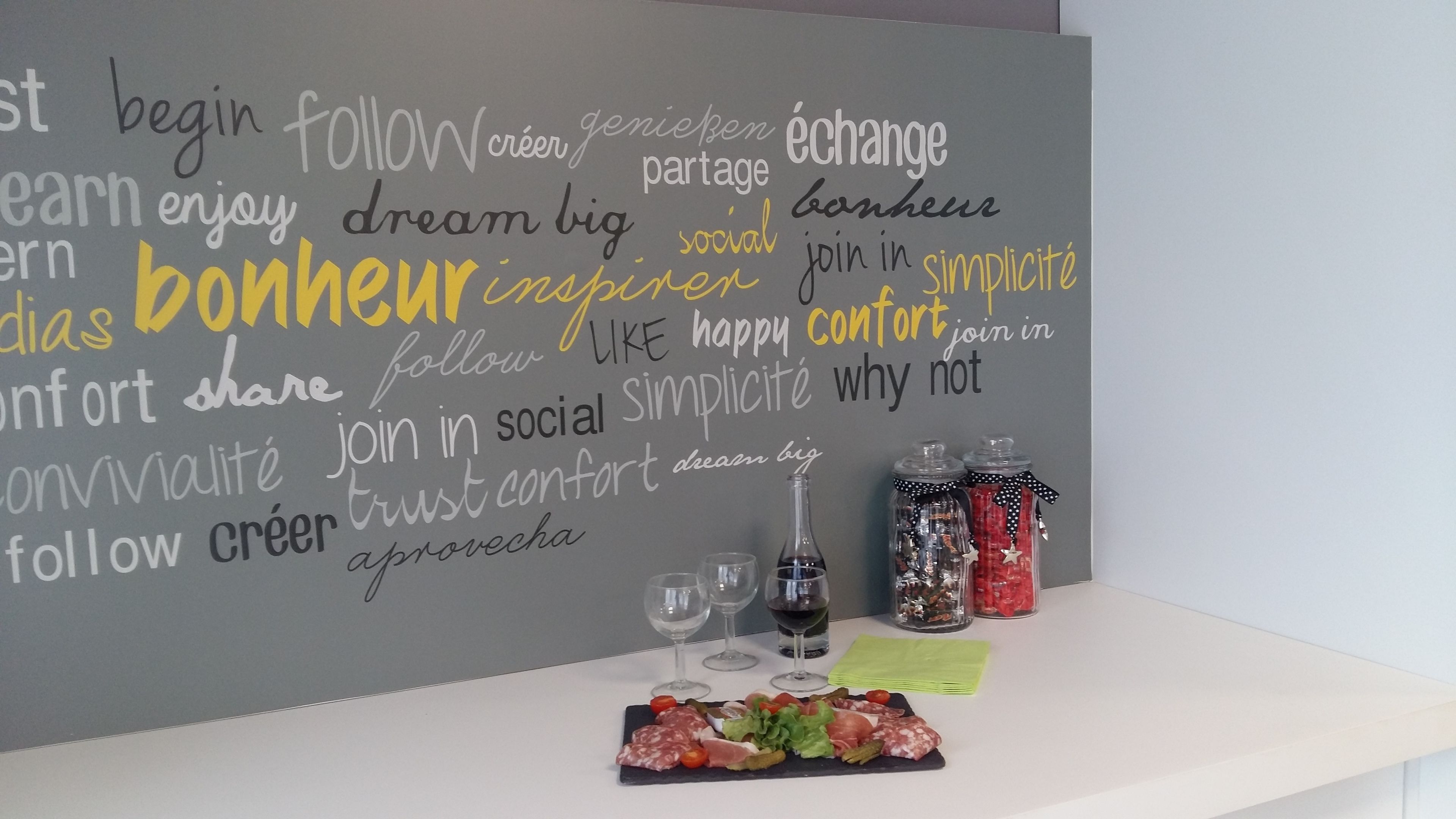 A wall with motivational words in multiple languages, a glass of red wine, two condiment jars, and a charcuterie board with various meats and vegetables.