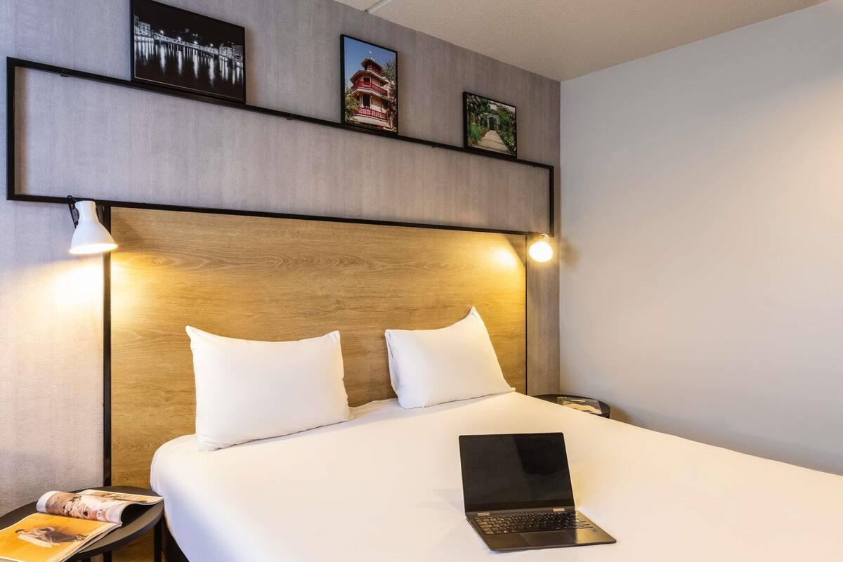 A hotel room with a large bed, two pillows, a laptop on the bed, a nightstand with a lamp, and framed pictures on the wall.