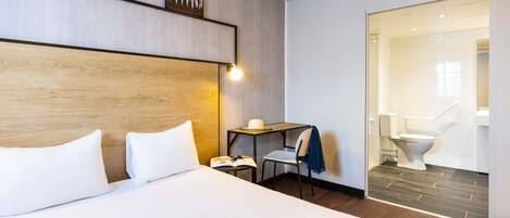 A hotel room with a bed, a desk, a chair, a nightstand, and a bathroom visible through an open door.