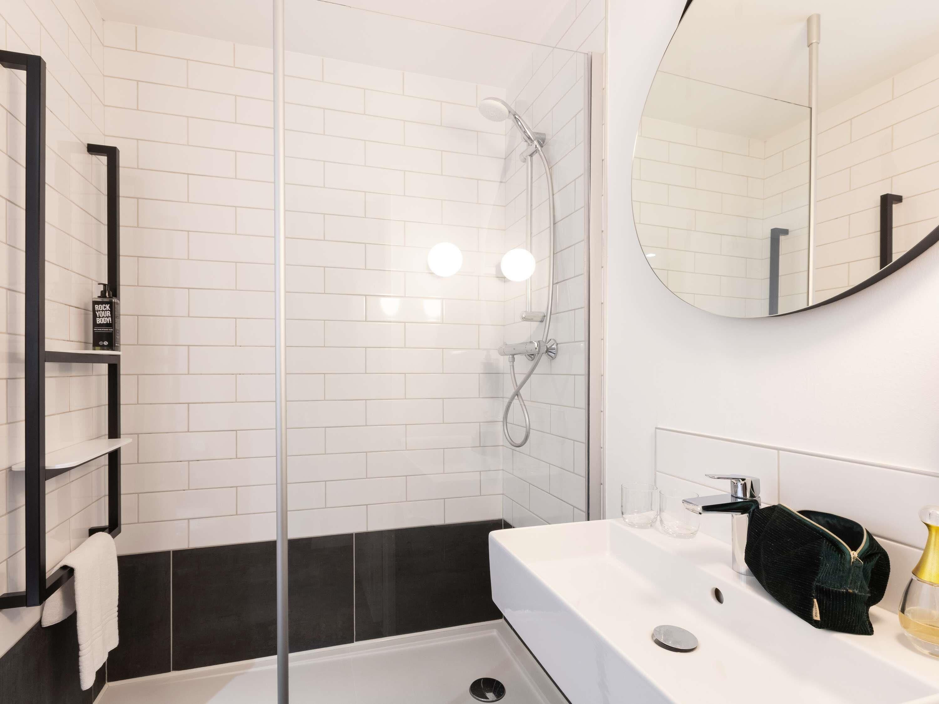 Une salle de bain moderne avec une douche à parois de verre, des carreaux blancs, un mur recouvert de carreaux noirs et blancs, un lavabo blanc avec une serviette foncée et un miroir rond.