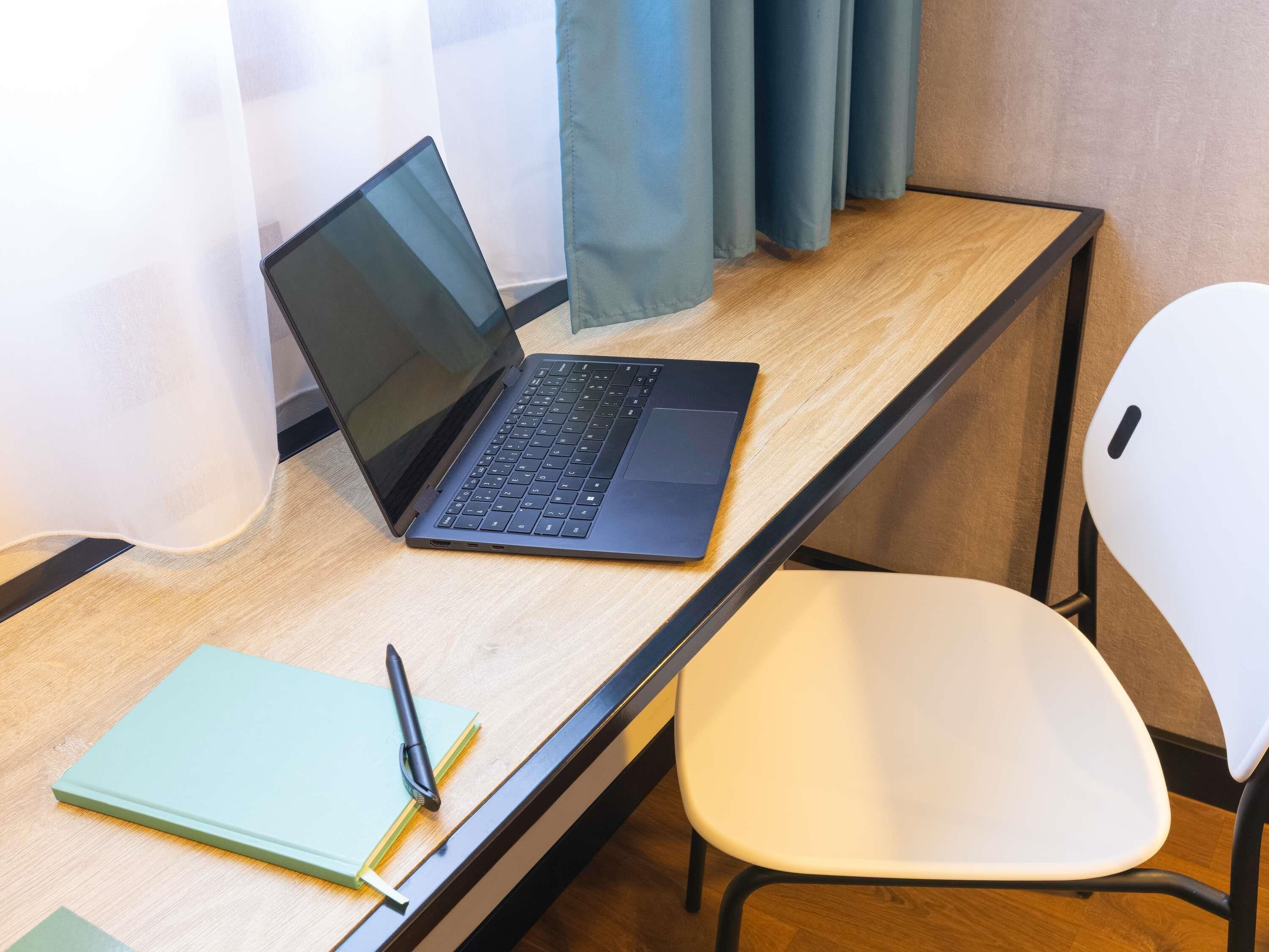 A laptop on a wooden desk with a white chair, a notebook, and a pen.