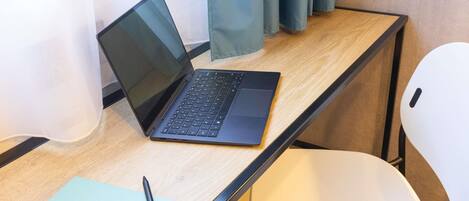 A laptop on a wooden desk with a white chair, a notebook, and a pen.