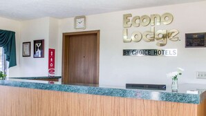 En hotelreception med en trædisk og et ur over disken. Væggen bag disken har et skilt, der står "Econo Lodge by Choice Hotels". Der hænger indrammede certifikater eller anerkendelser på væggen til venstre for døren.