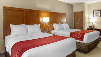 Habitación de hotel con dos camas, cada una con ropa de cama blanca y una manta roja. Tiene un cabecero de madera, una mesita de noche con una lámpara y un teléfono.