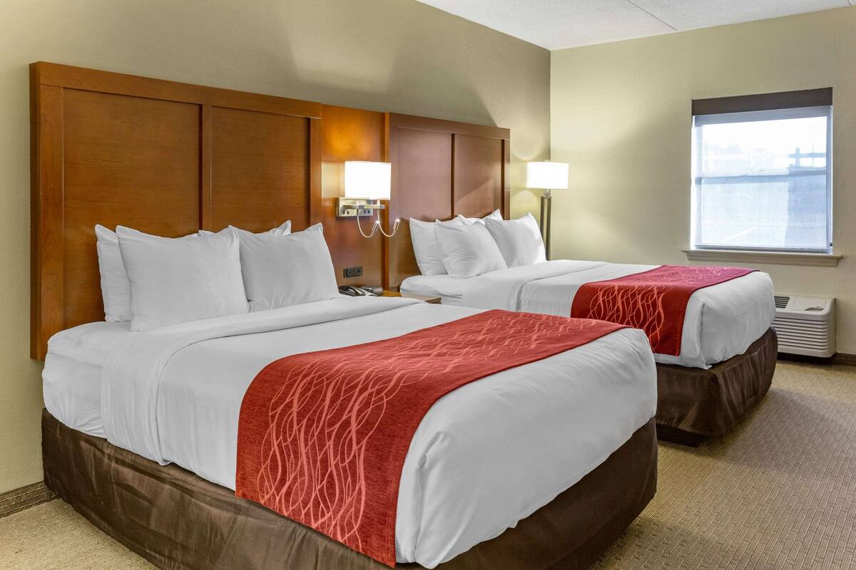 A hotel room with two beds, each with white linens and a red throw blanket. There are wall-mounted lamps and a window with a view.