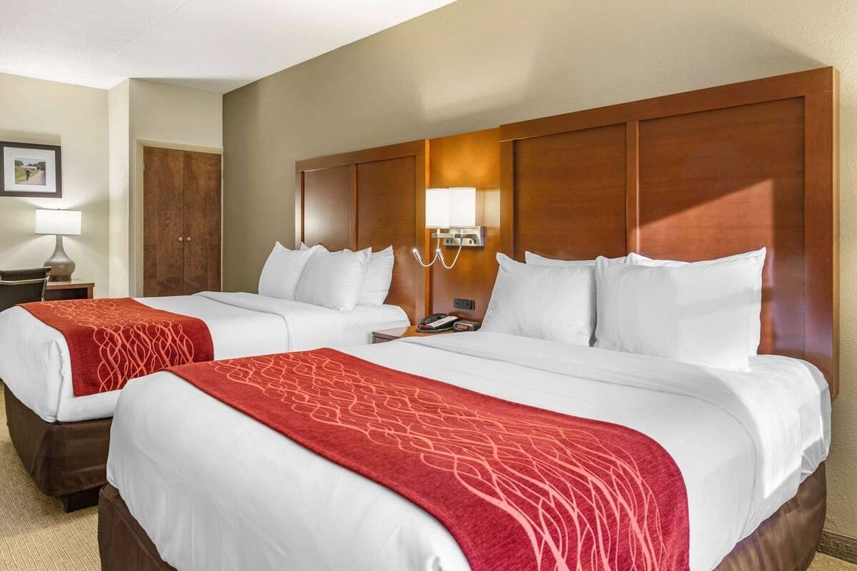 A hotel room with two beds, a nightstand with a lamp, a phone, and a wall-mounted light fixture.