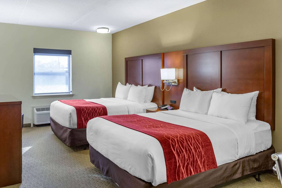 A hotel room with two beds, a desk, a lamp, and a window.