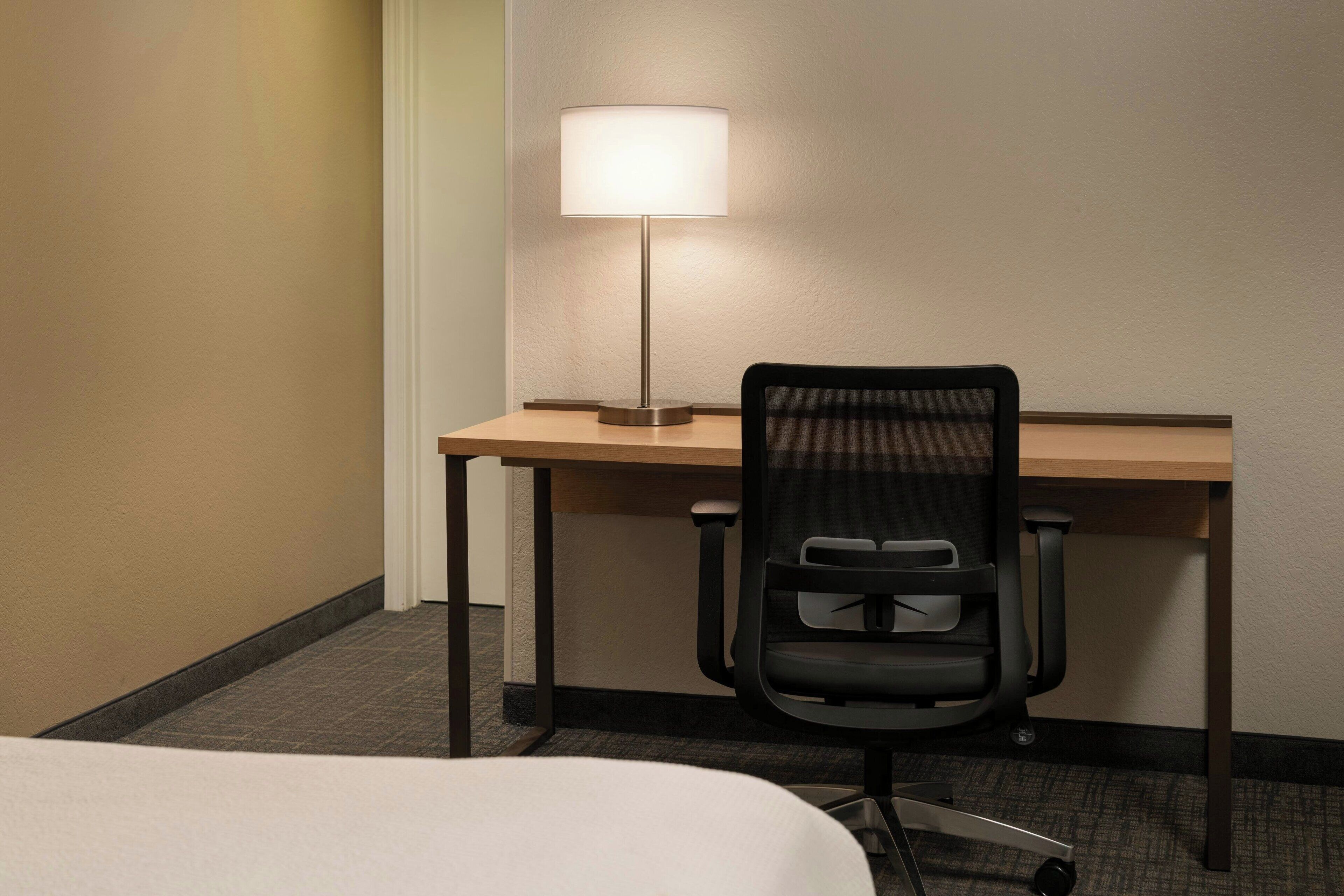 Een hotelkamer met een bed, een bureau met een lamp en een zwarte bureaustoel.
