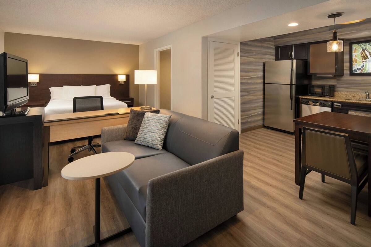 Une chambre d’hôtel moderne équipée d’un lit, d’un canapé, d’une petite table, d’une télévision, d’un bureau, d’une lampe, et d’un coin cuisine avec un réfrigérateur, un four à micro-ondes et un évier.