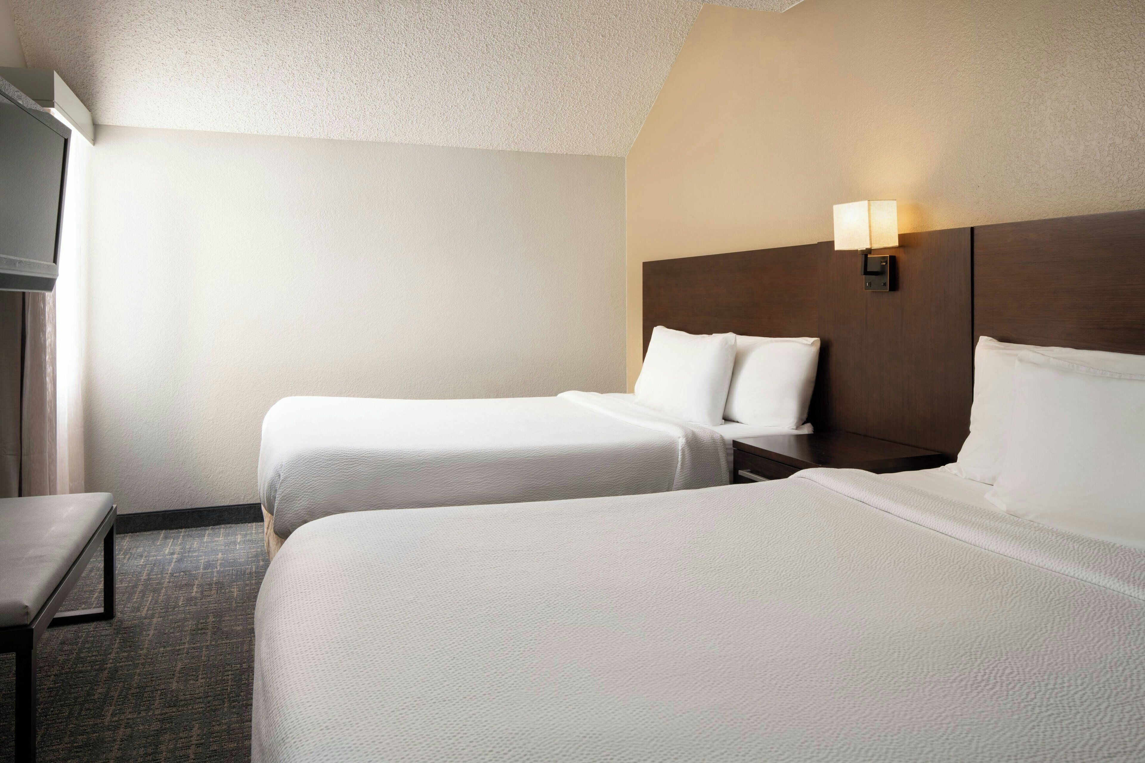 A hotel room with two beds, a nightstand, a lamp, and a window with curtains.