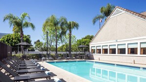 A pool area with lounge chairs and umbrellas, surrounded by palm trees and a building with large windows. - Residence Inn by Marriott Anaheim Placentia Fullerton (Placentia)