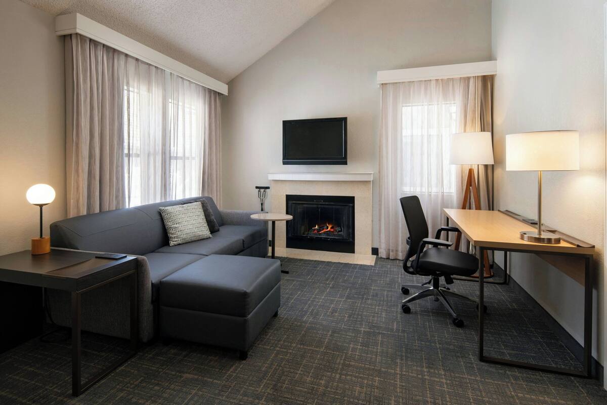 Une chambre d’hôtel moderne équipée d’un canapé, d’un bureau, d’une chaise, d’un téléviseur à écran plat fixé au-dessus d’une cheminée, et d’une lampe sur le bureau.